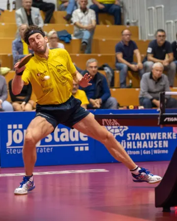 Spieler im gelben Trikot mit Stirnband im weiten Ausfallschritt am Tisch, dahinter Zuschauer auf der Tribüne