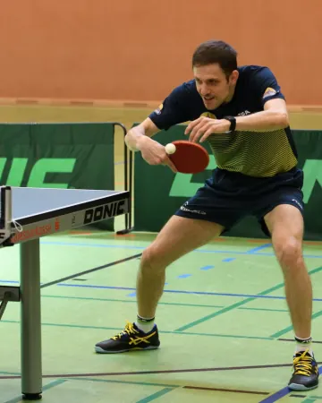 Spieler im dunkelblauen Trikot mit gelben Streifen wartet konzentriert auf den Ball, links ein Tisch mit Saalesparkasse-Aufdruck